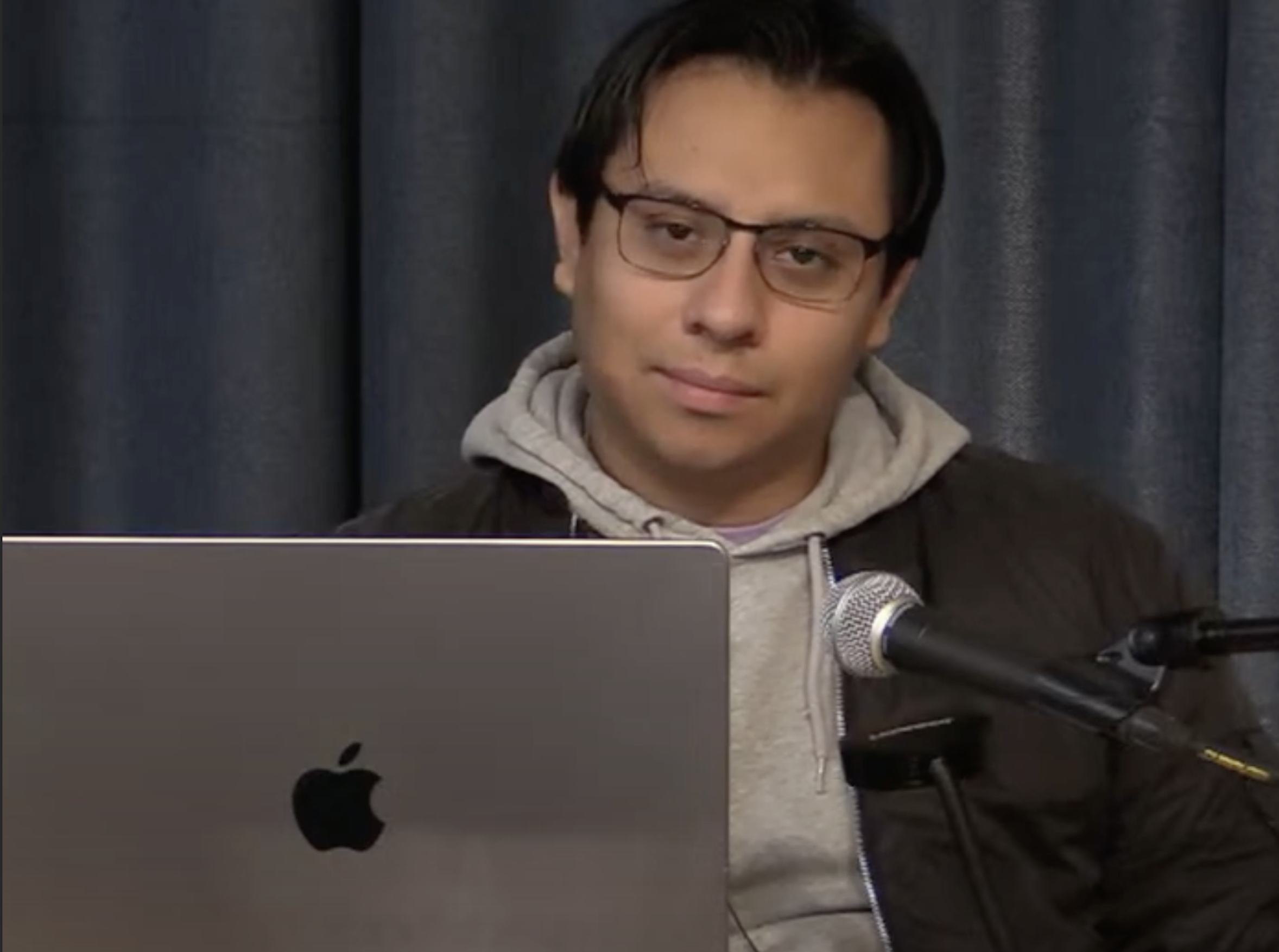 A person with short, dark hair and glasses is sitting in front of a dark curtain backdrop. They are wearing a gray hoodie under a dark jacket and looking slightly to the side with a neutral expression. In front of them is a laptop with an Apple logo. A microphone is positioned to the right of the laptop, suggesting that they might be speaking or preparing to speak.